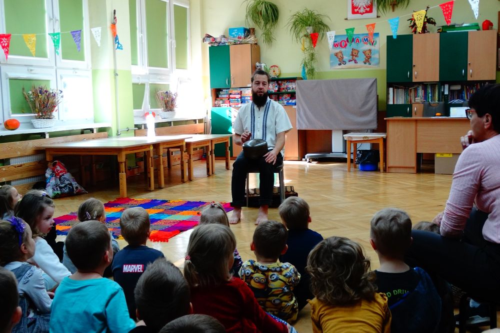 Pan Szymon Góralczyk siedzi przodem do dzieci z przedszkola, grając na istrumencie