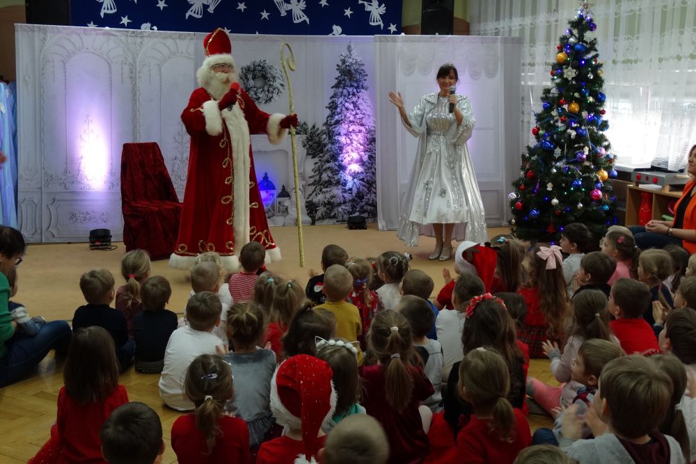 Święty Mikołaj i Mikołajowa (Artyści) stoją w rękach trzymając mikrofony. Dzieci siedzą na dywanie patrząc na artystów.