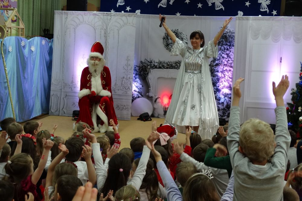 Mikołaj siedzi, Mikołajowa stoi z rękoma uniesionymi do góry. Dzieci siedzą na dywanie z rękoma uniesionymi do góry.