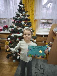 Dziewczynka stoi i trzyma w rękach złotą statuetkę i dyplom z wyróżnieniem. W tle jest choinka.