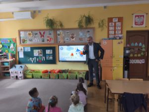 Dzieci siedzą na dywanie przodem do tablicy multimedialnej, na której wyświetlona jest prezentacja. Tata Ewy stoi przodem do dzieci i mówi