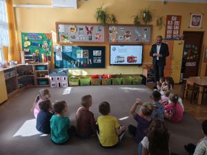 Dzieci siedzą na dywanie przodem do tablicy multimedialnej, na której wyświetlona jest prezentacja. Tata Ewy stoi przodem do dzieci i mówi