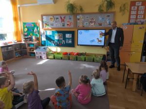 Dzieci siedzą na dywanie przodem do tablicy multimedialnej, na której wyświetlona jest prezentacja. Tata Ewy wskazuje ręką na dzieci które podnoszą ręce zgłaszając się do odpowiedzi.