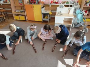 Dzieci siedzą na dywanie, przed każdym dzieckiem leżą kasztany ułożone w kolumnie