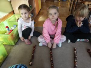 Dzieci siedzą na dywanie, przed każdym dzieckiem leżą kasztany ułożone w kolumnie