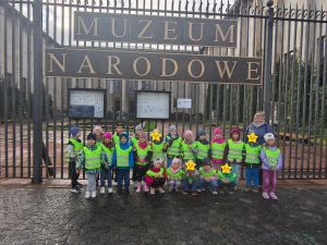 Zdjęcie grupowe. Dzieci stoją przed płotem muzeum, na którym jest napis MUZEUM NARODOWE