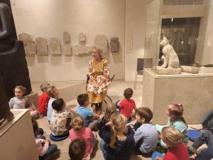 Dzieci siedzą na podłodze i słuchają edukatora muzealnego. Siedzą w sali w której są stare kamienie z Egiptu