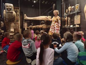 Dzieci siedzą na podłodze i słuchają edukatora muzealnego. W sali widać mumie i sarkofagi