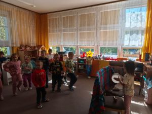 Dziewczynka stoi za parawanem z koca i gra na bębenku. A dzieci stoją na dywanie