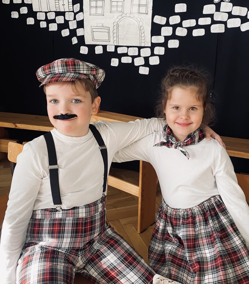 Chłopiec i dziewczynka siedzą na krzesłach, w tle dekoracja praskich kamienic. Dzieci ubrane w stroje dawnych prażan