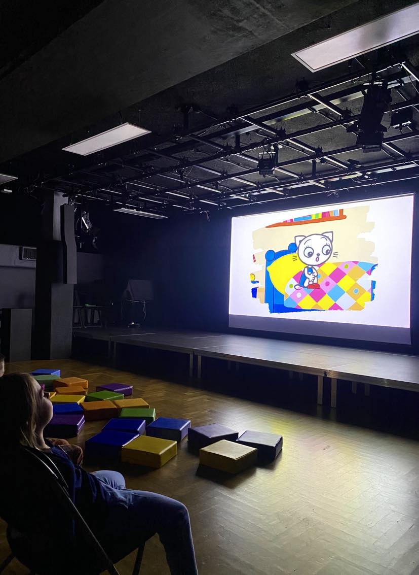 Dzieci siedzą na krzesłach i oglądają bajkę Kicia Kocia na dużym ekranie