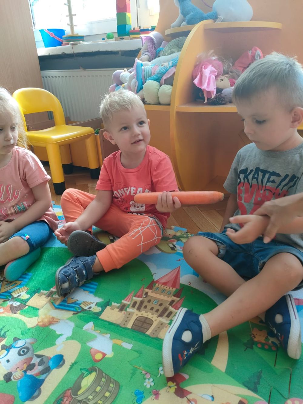 Dwóch chłopców siedzi na dywanie i podaje sobie marchewkę