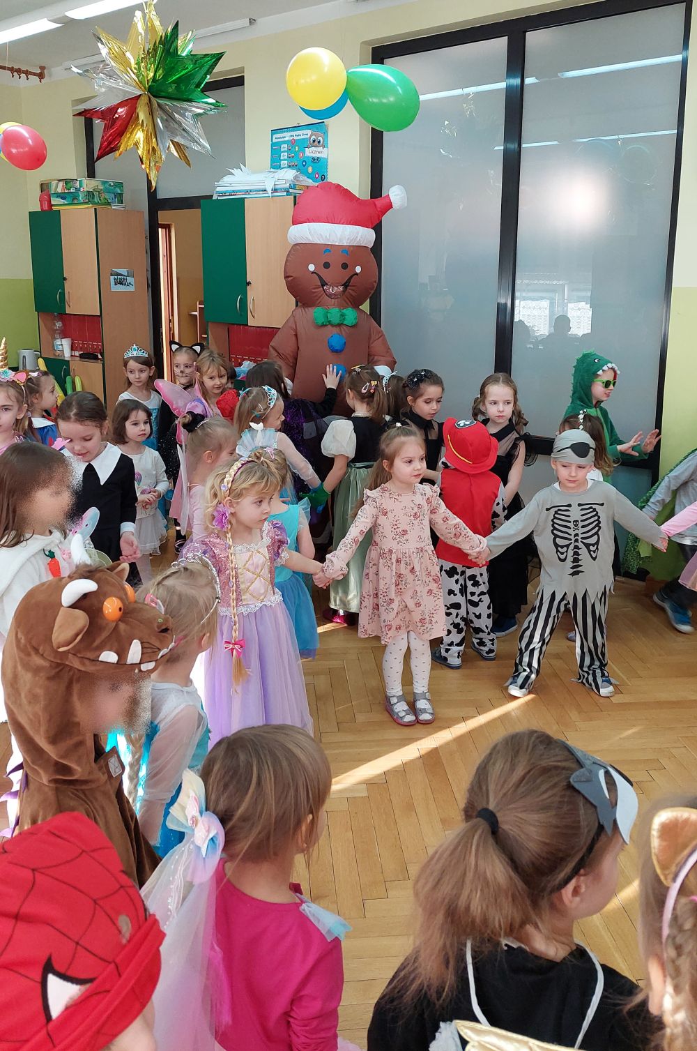 Dzieci ubrane w różne stroje tańczą w kole, z tyłu widać świątecznego pierniczka
