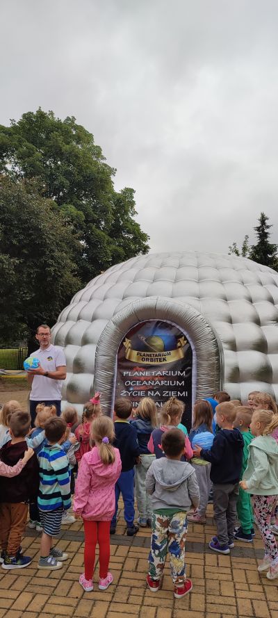 Dzieci stoją przed planetarium i słuchają Pana który opowiada o planetach