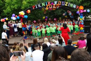 Na scenie dzieci z wszystkich grup śpiewają piosenkę, obok sceny widać stojących nauczycieli. Widać też publiczność