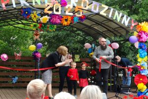Na zdjęciu przedszkolny amfiteatr ozdobiony balonami i kwiatami i napisem Piknik Rodzinny 2025. Na scenie stoją przedstawiciele władz miasta, Pani dyrektor i 2 dzieci z grupy biedronek. Pani i Pan z urzędu trzymają w rękach przeciętą czerwoną wstęgę