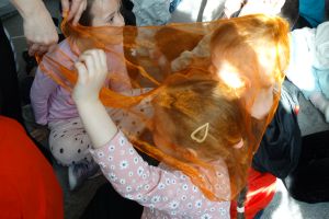 Kilkoro dzieci siedzi i jest przykryte przeźroczystą pomarańczową chustką